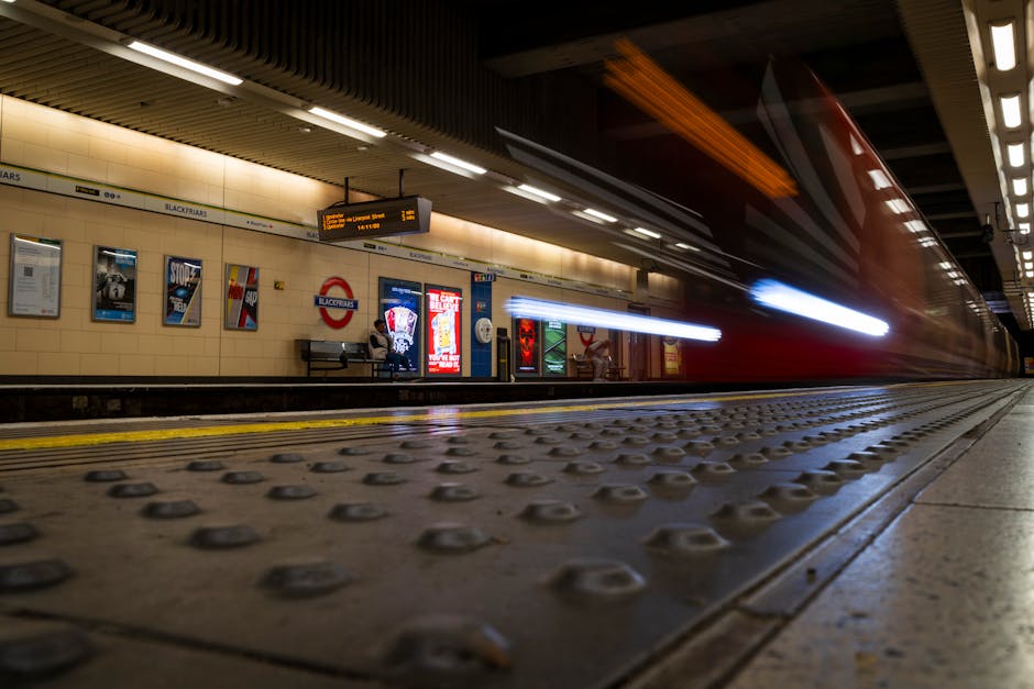 A blurred red train speeds through Colindale Tube station NW9, captured from a low angle on the platform showing textured tactile paving with rounded studs. The station wall in the background features