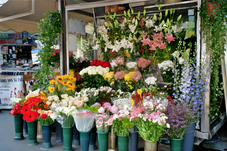 A vibrant flower stall featuring a diverse assortment of fresh floral arrangements and bouquets displayed outdoors. The composition includes tall white lilies with elongated buds and green stems, pink