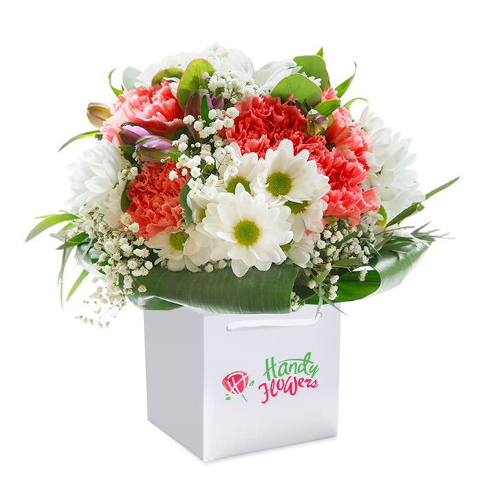 This striking bouquet, photographed with artistry against a stark white background, offers a sophisticated glimpse into Colindale's blend of tranquility and vibrancy. Clusters of coral and peach-hued carnations take center stage, their ruffled layers radiating with texture and warmth. Pops of brilliant red carnations infuse the composition with exuberance, while the interspersed white daisies-each with radiant yellow-green centers-bring a refreshing clarity, reminiscent of the open lawns at Rushgrove Park. Among the main blooms, tiny sprays of white baby's breath lend an ethereal softness, their fleeting quality echoing the gentle breezes through Colindale's shaded greenways. On the left, a few unopened buds with washes of lilac and purple hint at anticipation and new connections-much like the community's evolving charm. The bouquet is enveloped in lush, dark green foliage whose glossy leaves create a supportive, elegant wrap. Resting upright in a flawless white square gift bag with sturdy ribbon handles, the arrangement epitomizes ease of gifting and delivery throughout Colindale. Every detail speaks to the thoughtful craftsmanship of local florists, perfect for marking birthdays, anniversaries, or heartfelt moments across the area.
