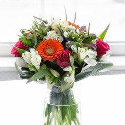 This masterfully arranged bouquet brings the beauty of Colindale's green spaces indoors, offering an exuberant celebration of colour and skilled design. Soft, creamy gerberas form a gentle, luminous wreath, their petals drawing the eye toward bold orange centres-reminiscent of sunset hues along the Grand Union Canal. Luxurious magenta roses anchor the arrangement with their rich tones, balanced by sprays of white lilies and filler blooms that cascade like spring blossoms through the streets of Colindale. Layered among the blooms are broad, glossy aspidistra leaves, shaped with care to cradle the collection and provide a structured, grounding presence. Delicate sprigs of green pierce the design, injecting freshness and organic movement. The bouquet is bathed in soft, natural light, lending each flower a vibrancy while highlighting subtle shadows and details. A gentle scent-rose mingled with green foliage-hovers above the vase, evoking the peaceful calm of Colindale's parks. Perfect for gatherings, housewarmings, or showing gratitude, this arrangement from skilled Colindale florists captures hope, celebration, and a deep connection to local flora and community spirit.